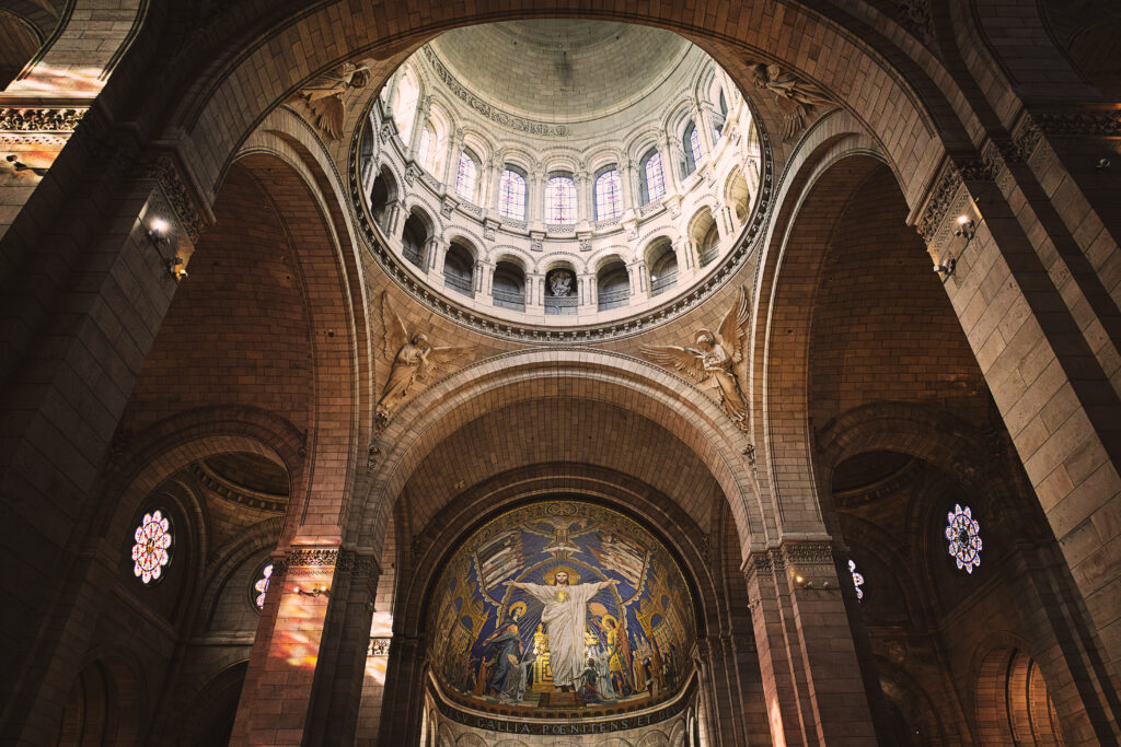 Photographie en couleur de la coupole du Sacré-Coeur à Paris. Au centre, en haut, la coupole soutenue par des voutes crée un puits de lumière éclairant le reste de la photographie. Au centre, en dessous de la coupole, quatre anges tenant chacun dans leurs main les instruments de la Passion du Christ. Au centre, en bas, se situe la grande mosaïque montrant le Christ, bras ouverts, divers figures historiques dont Jeanne d'Arc, et personnages ayant contribué à l'histoire Chrétienne ou de la basilique.