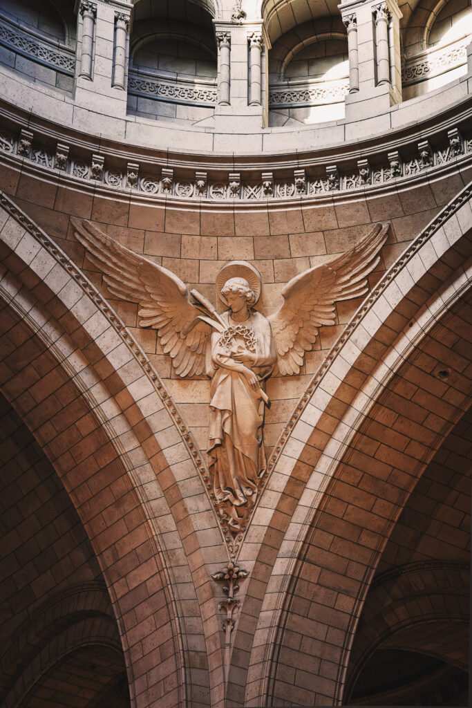 Photographie en couleur d'un des anges du Sacré Cœur de Paris. Celui pris porte dans ses bras la couronne de la passion du Christ contre son cœur. Ses ailes son déployées, il regarde vers le bas-gauche. Au dessus de lui plusieurs voutes sont visibles, ils soutiennent la coupole plus haut qui n'est pas visible sur la photographie.
