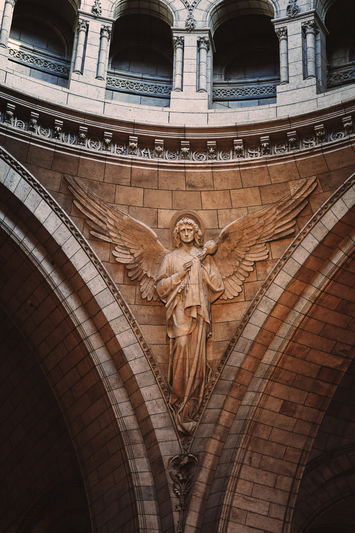Photographie en couleur d'un des anges du Sacré Cœur de Paris. Celui pris porte sur sa poitrine une éponge imbibée de vinaigre, l'un des instruments de la Passion. Ses ailes sont déployées, il regarde vers nous. Au dessus de lui plusieurs voutes sont visibles, ils soutiennent la coupole plus haut qui n'est pas visible sur la photographie.