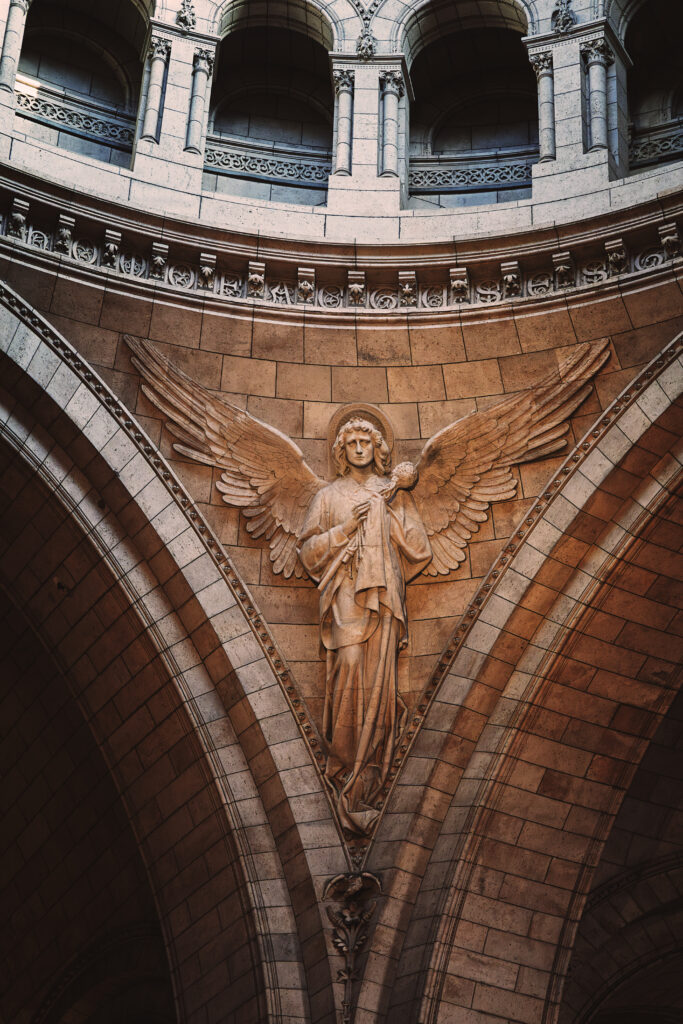 Photographie en couleur d'un des anges du Sacré Cœur de Paris. Celui pris porte sur sa poitrine une éponge imbibée de vinaigre, l'un des instruments de la Passion. Ses ailes sont déployées, il regarde vers nous. Au dessus de lui plusieurs voutes sont visibles, ils soutiennent la coupole plus haut qui n'est pas visible sur la photographie.