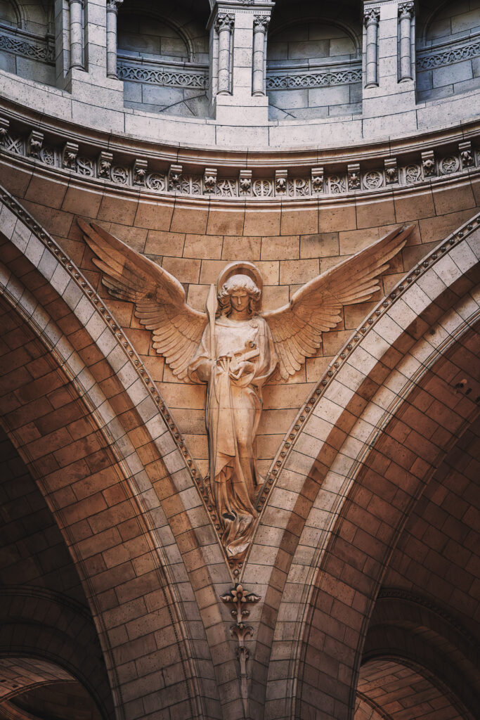 Photographie en couleur d'un des anges du Sacré Cœur de Paris. Celui pris tient dans sa main gauche la lance et dans sa main droite les clous ayant servi à crucifier le Christ. Ses ailes sont déployées, il regarde vers le bas. Au dessus de lui plusieurs voutes sont visibles, ils soutiennent la coupole plus haut qui n'est pas visible sur la photographie.
