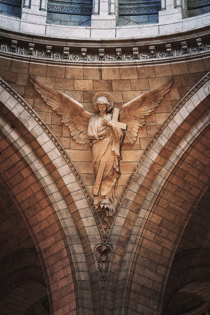 Photographie en couleur d'un des anges du Sacré Cœur de Paris. Celui pris tient contre lui la sainte croix. Ses ailes sont déployées, il regarde vers le bas. Au dessus de lui plusieurs voutes sont visibles, ils soutiennent la coupole plus haut qui n'est pas visible sur la photographie.