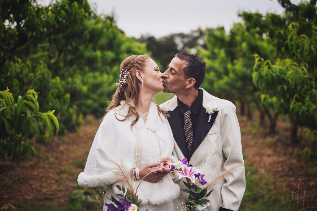 Photographie en couleur d'un couple de mariés s'embrassant au milieu d'un verger.