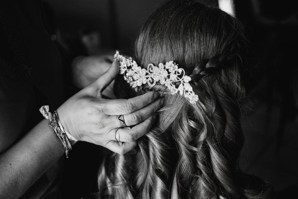 Photographie en noir et blanc de la préparation d'une coiffure pour un mariage. On y voit la main de la coiffeuse tenir une barrette décorative dans les cheveux de la marié, qui ondulent en anglaise juste dessous.