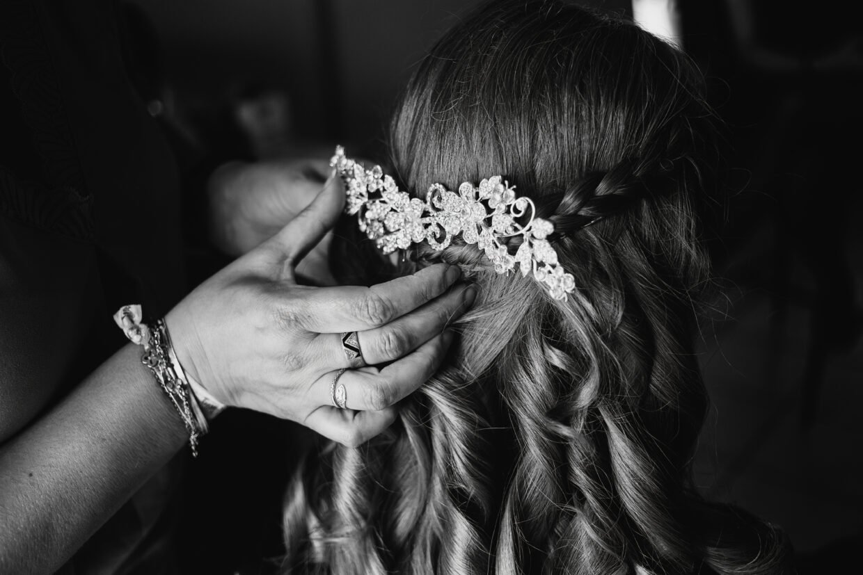 Photographie en noir et blanc de la préparation d'une coiffure pour un mariage. On y voit la main de la coiffeuse tenir une barrette décorative dans les cheveux de la marié, qui ondulent en anglaise juste dessous.
