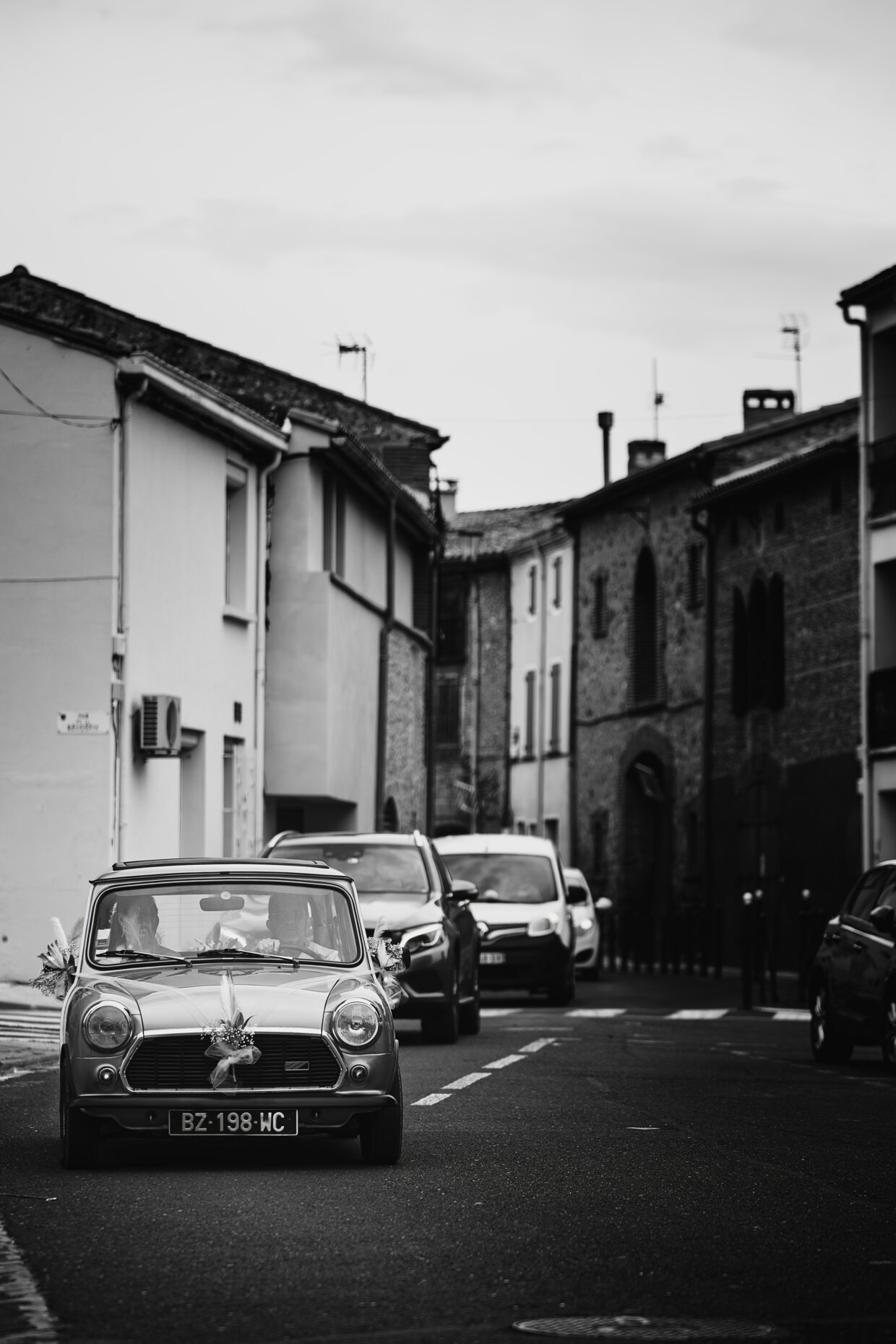 Photographie en noir et blanc dans la rue. Elle dépeint l'arrivée de la voiture d'une mariée à la mairie. En premier plan la voiture, très ancienne, dont je n'ai malheureusement pas le modèle. Derrière elle plusieurs autres voitures plus modernes la suive. Des maisons bordant la route sont visibles au second plan.