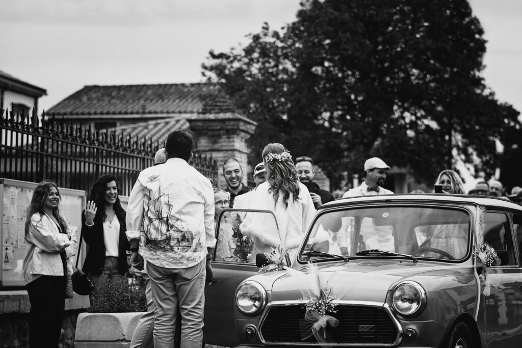 Photographie en noir et blanc montrant l'arrivée d'une mariée à la mairie. Elle vient de descendre de sa voiture et fait face à ses invités. Les personnes sourient et sont visiblement très joyeuses.