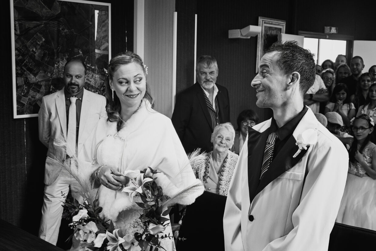 Photographie en noir et blanc de deux mariés sur le point de se dire "oui" à la mairie. Au premier plan, à gauche, la mariée en train de regarder son compagnon avec un regard amusé et un grand sourire. A droite, l’époux, lui rendant son regard amusé. Dans le fond, les invités attendent et semble suspendu à la réponse.