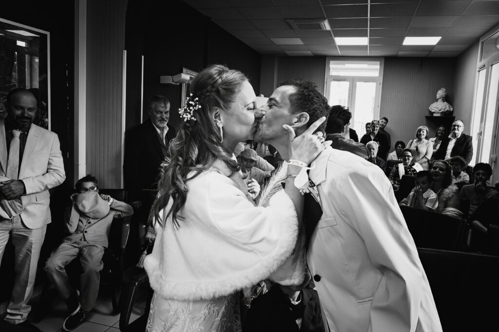 Photographie en noir et blanc d'un couple de marié en train de s'embrasser, souriants. la mariée est à gauche, son époux est à droite. Dans le fond les invités sourient en les regardant.