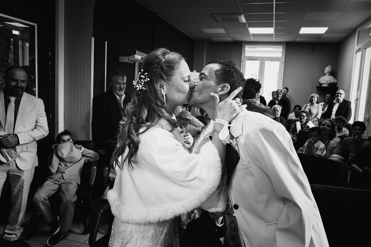 Photographie en noir et blanc d'un couple de marié en train de s'embrasser, souriants. la mariée est à gauche, son époux est à droite. Dans le fond les invités sourient en les regardant.