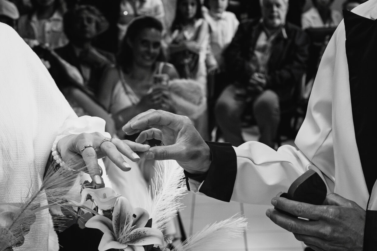 Photographie en noir et blanc de l'échange des anneaux lors d'un mariage. On y voit en premier plan les mains des mariés. A droite, celle de la mariée. Juste à côté la main de l'époux s'éloigne, juste après avoir glissé l'anneau au doigt de sa compagne. En second plan, les invités, souriant.