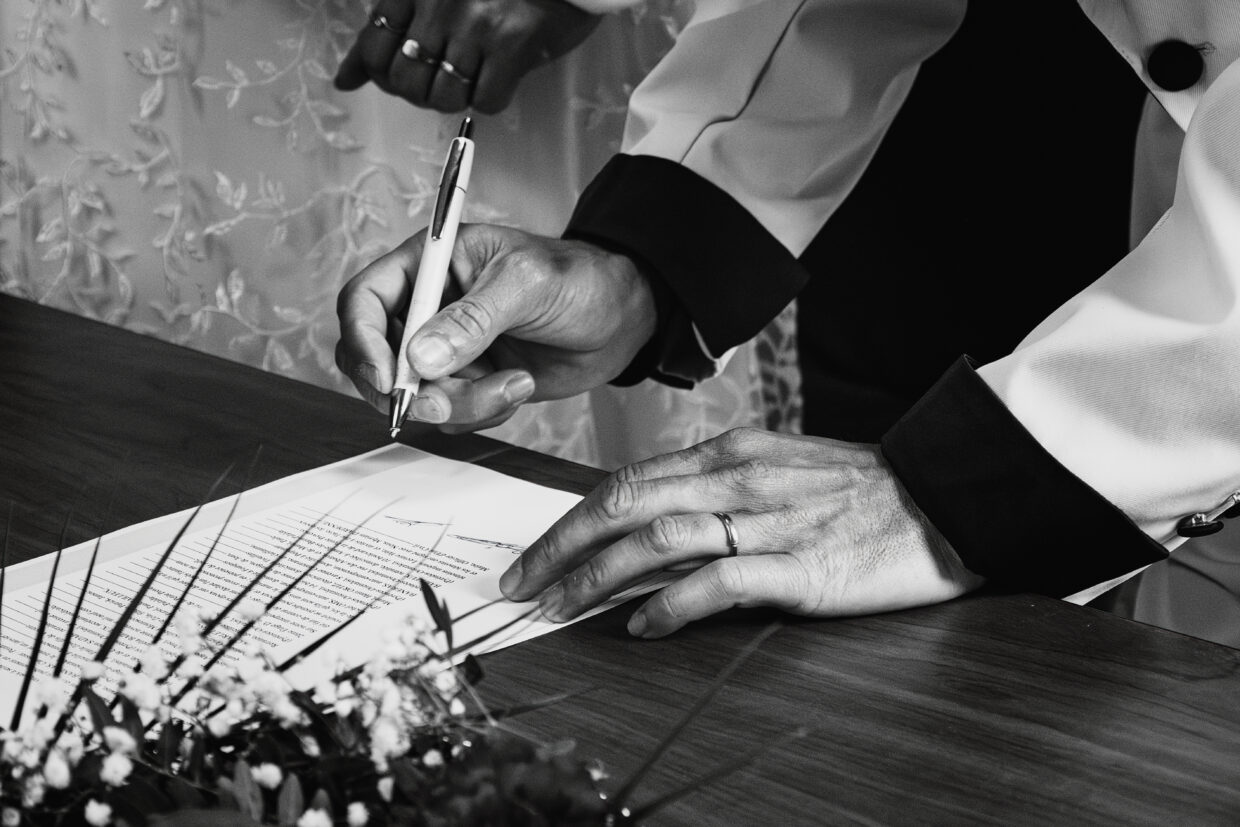 Photographie en noir et blanc de la signature des actes de mariage. Ici le marié est en train de signer. La photo est cadrée sur ses mains, le stylo est légèrement au dessus de la feuille.
