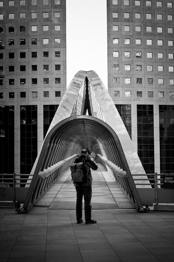 Photographie en noir et blanc d'un pont couvert à la Défense. Devant, au centre, un photographe est visible, en train de prendre en photo des personnes en train de traverser à l'intérieur du pont-tunnel.