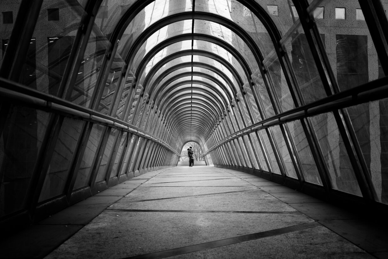 Photographie en noir et blanc de l'intérieur d'un pont-tunnel se situant à la Défense à Paris. La photographie est très géométrique et graphique, amenant le regard vers le centre, où tout au bout, à l'entrée, se trouvent deux personnes.