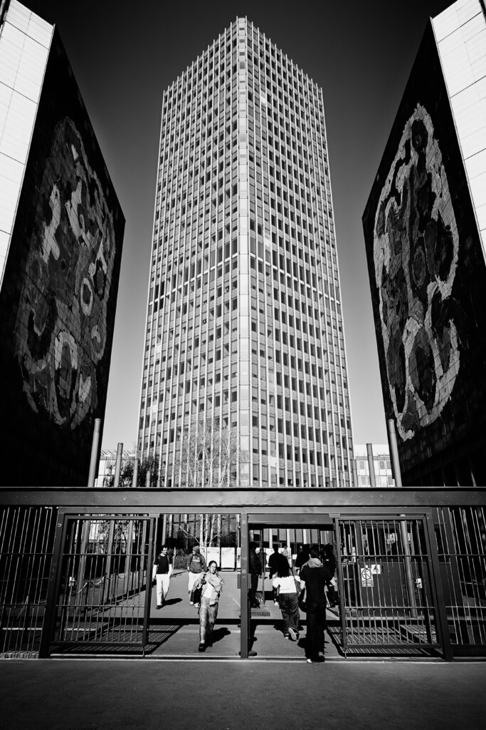 Photographie en noir et blanc de l'entrée d'une université à Paris (laquelle, par contre, bonne question, une extension de la Sorbonne peut être ?). Sur le bas de la photo une grille avec des portes ouvertes permettent à des jeunes personnes d'entrer et sortir. En fond, un bâtiment extrêmement haut, entouré, au premier plan, par deux autres immeubles aux murs peints. La photo est très symétrique.