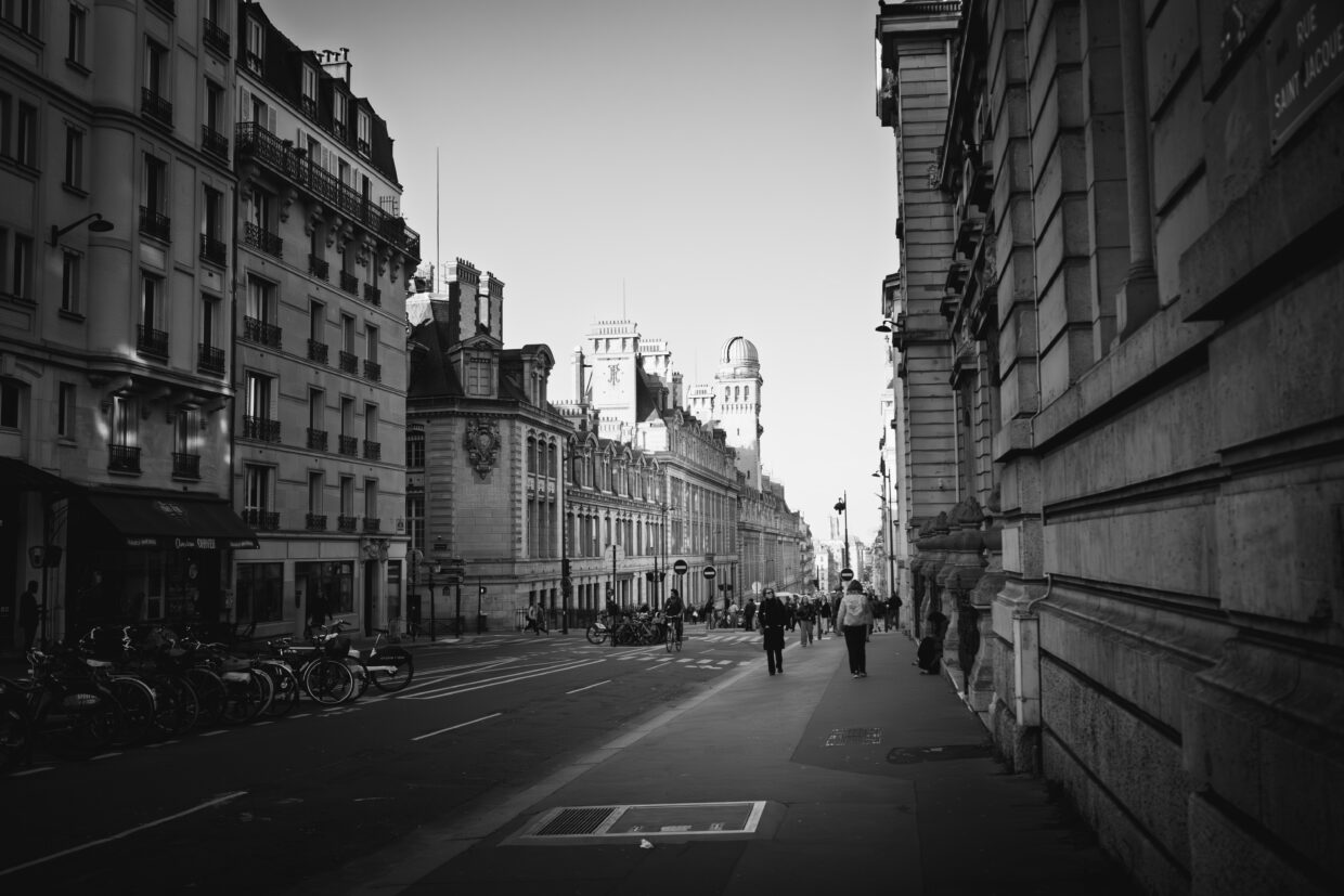 Photographie en noir et blanc de la rue Saint Jacques, à Paris, proche du Panthéon. De hauts immeubles haussmanniens bordent la rue. Aucune voiture n'est visible, mais de nombreux deux roues sont garés à gauche de la photo. Vers le point de fuite des piétons marchent sur le trottoir.