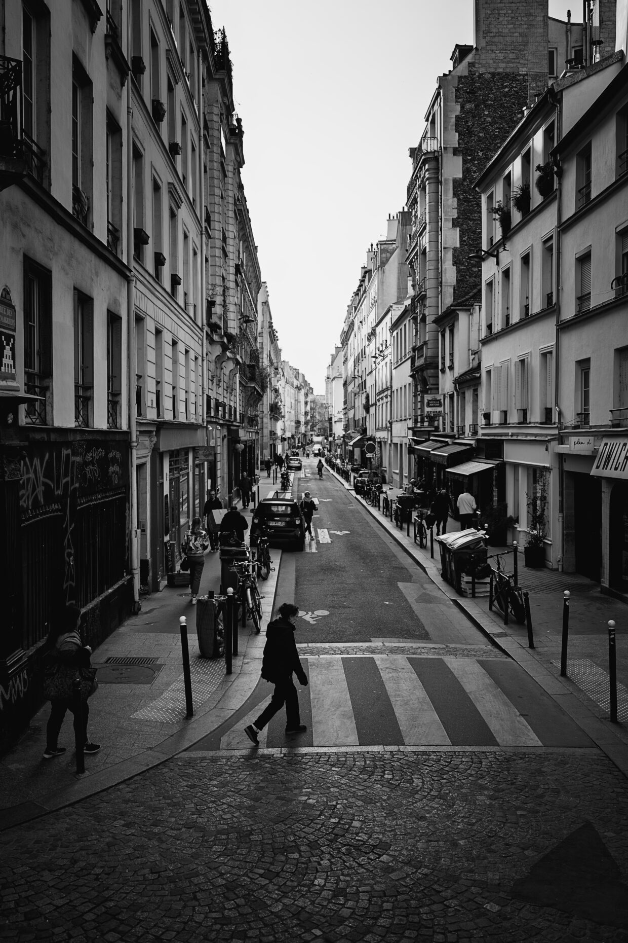 Photographie en noir et blanc de la rue des Vinaigriers, dans le dixième arrondissement de Paris. De chaque côté d'une route des immeubles, de style et époques différentes, bordent une petite route transformée en piste cyclable. Sur les trottoirs plusieurs piétons marchent. Des voitures sont également garées. En bas, sur un passage piéton, une personne traverse.