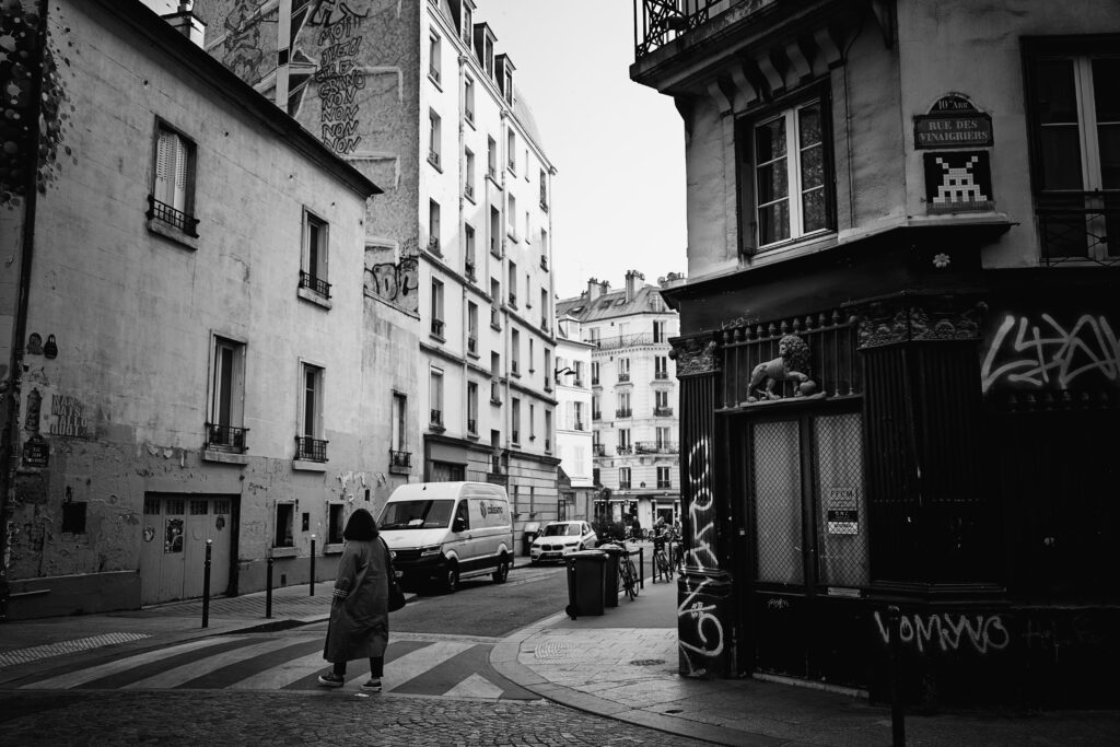Photographie en noir et blanc de l'angle de la rue des vinaigriers, dans le 10ème arrondissement de Paris. A droite, une porte grillagée est surmontée de la statue d'un lion avec une patte posée sur une sphère. Un peu plus haut, sous le panneau de la rue, une mosaïque street art représentant un space invader est visible. A gauche, traversant un passage piéton, une personne est visible de dos. le reste de la photo est composée de bâtiment de plusieurs étages, bordant les routes.