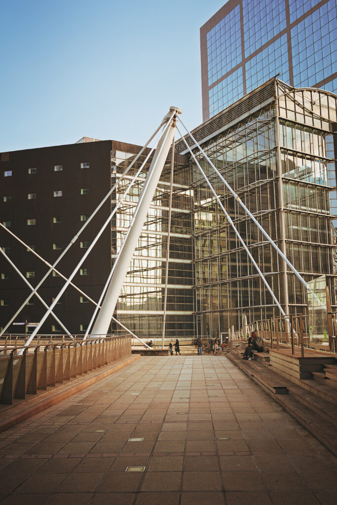 Photographie de rue d'une passerelle de la Défense, proche de l'arche bien connue. On y voit au centre une structure en métal blanc triangulaire qui soutient probablement la passerelle. Dans le fond plusieurs bâtiments, certains entièrement recouverts de fenêtres. Assis sur des bancs disposés sur la passerelle sont présents plusieurs personnes. D'autres marchent plus loin.