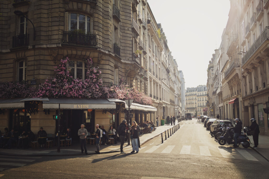 Photographie de rue, à Paris, sur laquelle figure la devanture du café La Marine, devant laquelle plusieurs personnes sont attablées, ou discutent debout sur la chaussée. Le premier étage du bâtiment haussmannien est recouvert de fleurs rose. Sur la partie droite de la photo, une rue bordée d'immeubles du même type fait un point de fuite.
