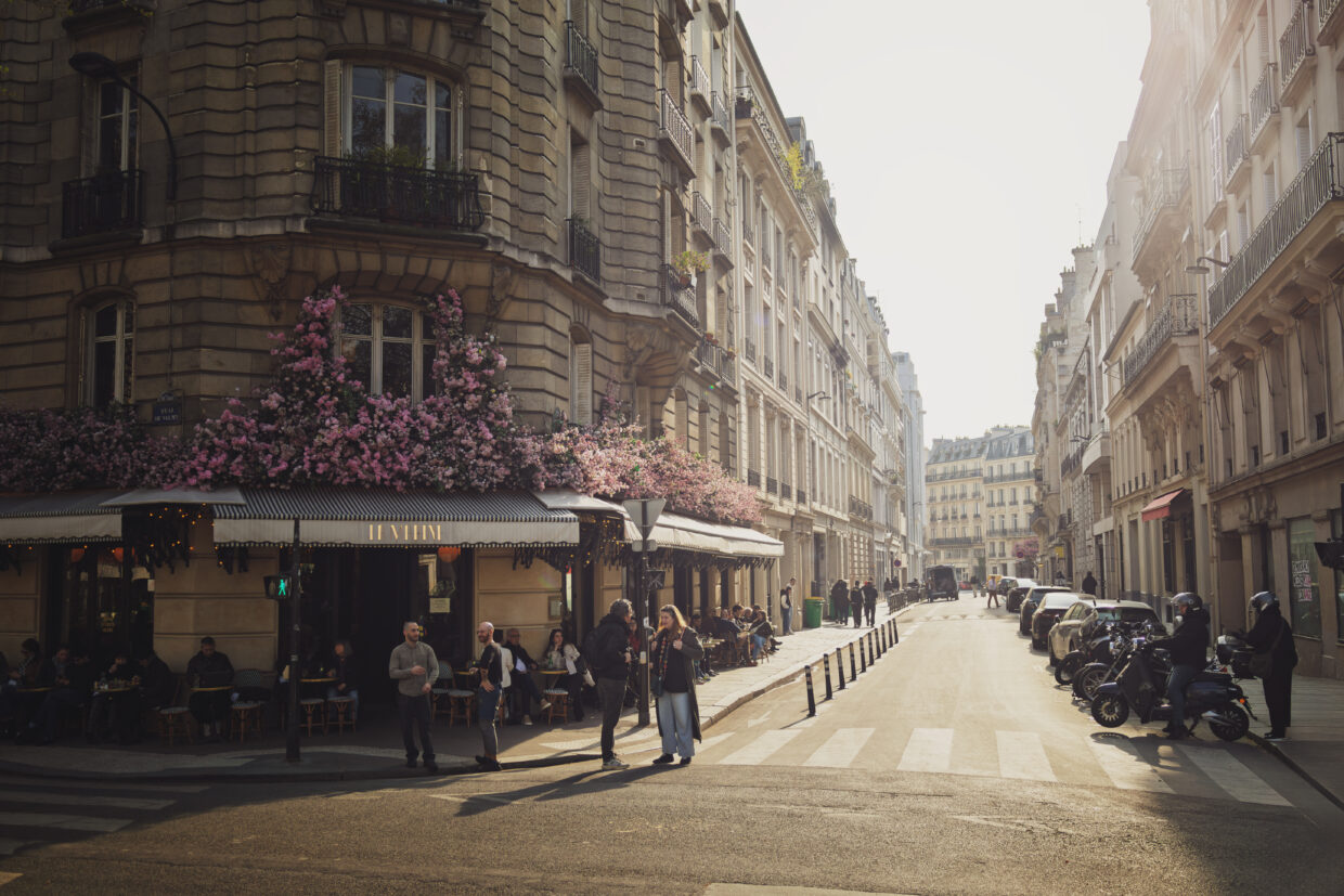 Photographie de rue, à Paris, sur laquelle figure la devanture du café La Marine, devant laquelle plusieurs personnes sont attablées, ou discutent debout sur la chaussée. Le premier étage du bâtiment haussmannien est recouvert de fleurs rose. Sur la partie droite de la photo, une rue bordée d'immeubles du même type fait un point de fuite.