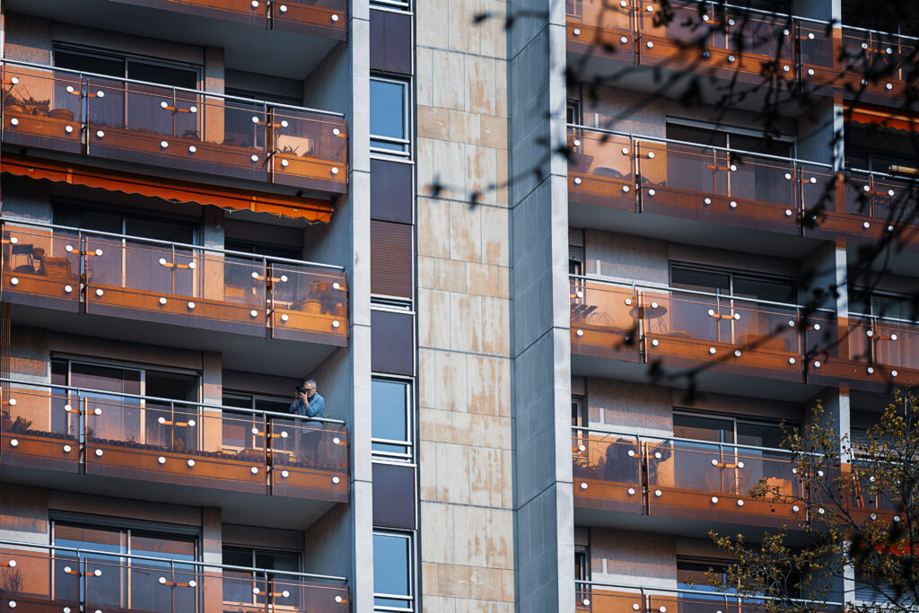 Photographie en couleur d'une façade d'un bâtiment d'habitations, composé de nombreux balcons. Sur l'un d'eux, en bas à gauche, un photographe est en train de prendre des photos. Le bâtiment était assez loin de mon point de vue et le photographe se trouvait au mois au quatrième ou cinquième étage. L'idée était donc de prendre en photo le photographe qui se pensait à l'abri :D