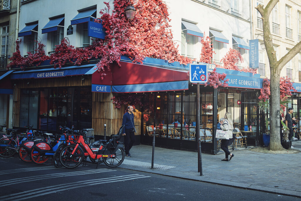 Photographie de rue d'une vitrine de boulangerie sur laquelle est écrit "Pain Bio au levain". La mise au point est faite sur le reflet des bâtiments et terrasse qui se trouve en face de la boulangerie. Le double vitrage cause un effet de dédoublement, comme si deux images n'étaient pas parfaitement alignée. Cet effet est renforcé par l'intérieur de la boulangerie qui est visible, particulièrement en bas de la vitrine, ajoutant un niveau supplémentaire aux images qui se mélangent.