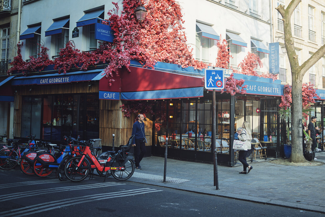 Photographie de rue d'une vitrine de boulangerie sur laquelle est écrit "Pain Bio au levain". La mise au point est faite sur le reflet des bâtiments et terrasse qui se trouve en face de la boulangerie. Le double vitrage cause un effet de dédoublement, comme si deux images n'étaient pas parfaitement alignée. Cet effet est renforcé par l'intérieur de la boulangerie qui est visible, particulièrement en bas de la vitrine, ajoutant un niveau supplémentaire aux images qui se mélangent.