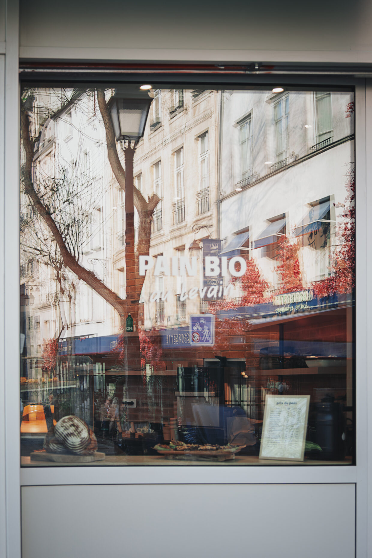 Photographie de rue d'une vitrine de boulangerie sur laquelle est écrit "Pain Bio au levain". La mise au point est faite sur le reflet des bâtiments et terrasse qui se trouve en face de la boulangerie. Le double vitrage cause un effet de dédoublement, comme si deux images n'étaient pas parfaitement alignée. Cet effet est renforcé par l'intérieur de la boulangerie qui est visible, particulièrement en bas de la vitrine, ajoutant un niveau supplémentaire aux images qui se mélangent.