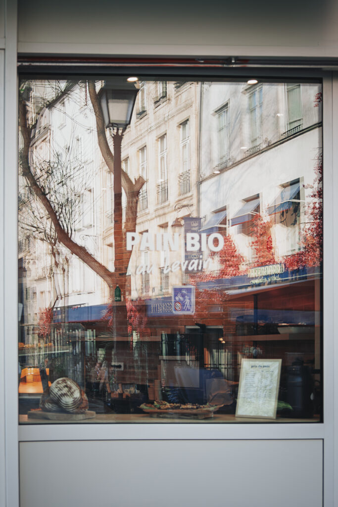 Photographie de rue d'une vitrine de boulangerie sur laquelle est écrit "Pain Bio au levain". La mise au point est faite sur le reflet des bâtiments et terrasse qui se trouve en face de la boulangerie. Le double vitrage cause un effet de dédoublement, comme si deux images n'étaient pas parfaitement alignée. Cet effet est renforcé par l'intérieur de la boulangerie qui est visible, particulièrement en bas de la vitrine, ajoutant un niveau supplémentaire aux images qui se mélangent.