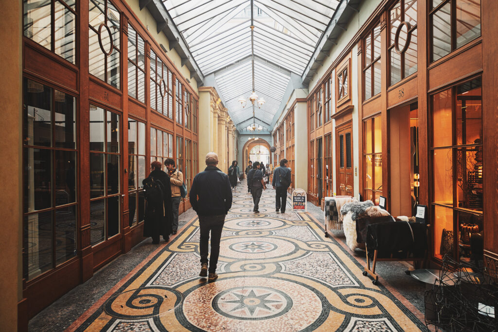 Photographie de l'intérieur de la galerie Vivienne à Paris. De part et d'autre de l'allée pavée de mosaïque des devantures faites de bois et de vitre montre l'intérieur de plusieurs boutiques et café-restaurants. Plusieurs personnes marchent le long et regardent l'intérieur des vitrines. Le plafond est entièrement constituées de verre fumé, laissant ainsi la lumière naturelle éclairer la galerie.